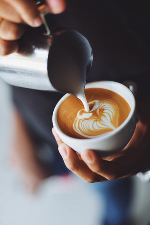 a hand holding a cup while the other hold a metal jug the metal jug is pouring milk into the cup with espresso in it creating latte art in the shape of a heart